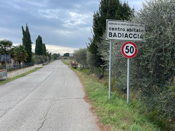 Section de piste cyclable sur une route de campagne asphaltée avec, à droite, un panneau indiquant l'entrée du village de Badiaccia